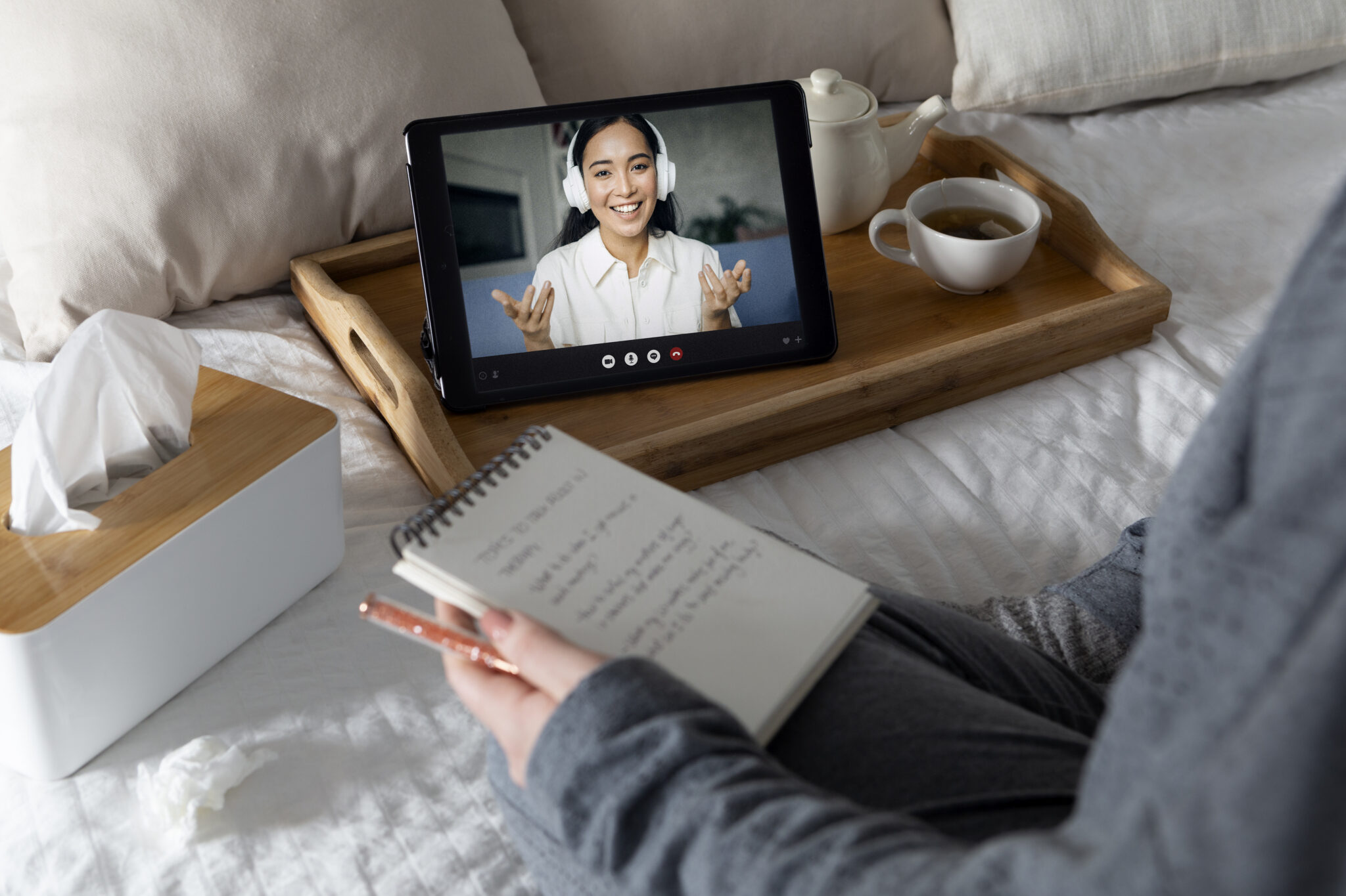 Widoczny profil osoby siedzącej na łóżku. W rękach trzyma notatnik i długopis. Na łóżku pudełko chusteczek i taca z filiżanką herbaty i dzbankiem. Obok filiżanki tablet, na którym widoczna jest uśmiechnięta osoba ubrana na biało, ze słuchawkami na uszach. Żywo gestykuluje. Ilustracja do obrazka Po co dorosłej osobie diagnoza ADHD?