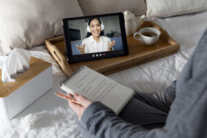 Najważniejszy wywiad o ADHD w moim życiu 2 Widoczny profil osoby siedzącej na łóżku. W rękach trzyma notatnik i długopis. Na łóżku pudełko chusteczek i taca z filiżanką herbaty i dzbankiem. Obok filiżanki tablet, na którym widoczna jest uśmiechnięta osoba ubrana na biało, ze słuchawkami na uszach. Żywo gestykuluje. Ilustracja do obrazka Po co dorosłej osobie diagnoza ADHD?